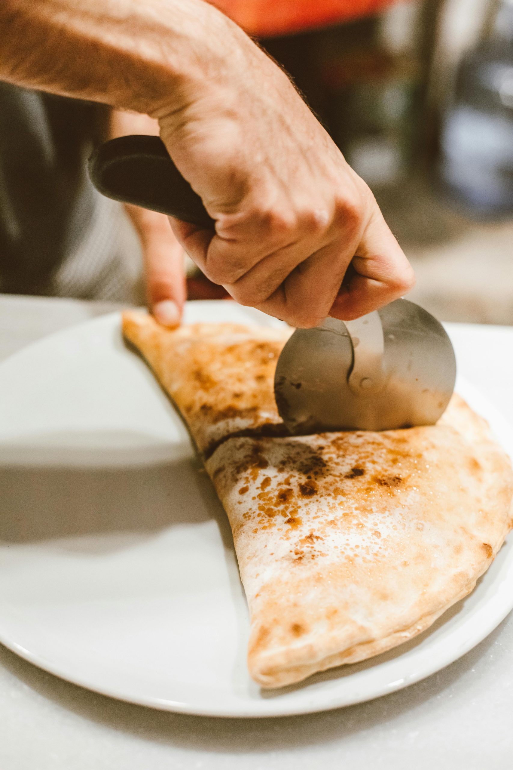 Hand slicing calzone with a pizza cutter on a white plate, showcasing food preparation.